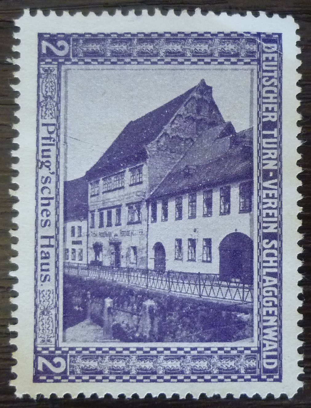 Reklamní/spolková nálepka (vinětka) vydaná spolkem Deutscher Turn-Verein Schlaggenwald (Německý tělocvičný spolek Schlaggenwald), s vyobrazením domu zvaného „Pflug'sches Haus" (Pflugův dům) v Horním Slavkově – patrový roubený/zděný dům se štítem u potoka s můstkem a zábradlím. Na nálepce nápisy: „DEUTSCHER TURN-VEREIN SCHLAGGENWALD", „Pflug'sches Haus." a hodnota „2".