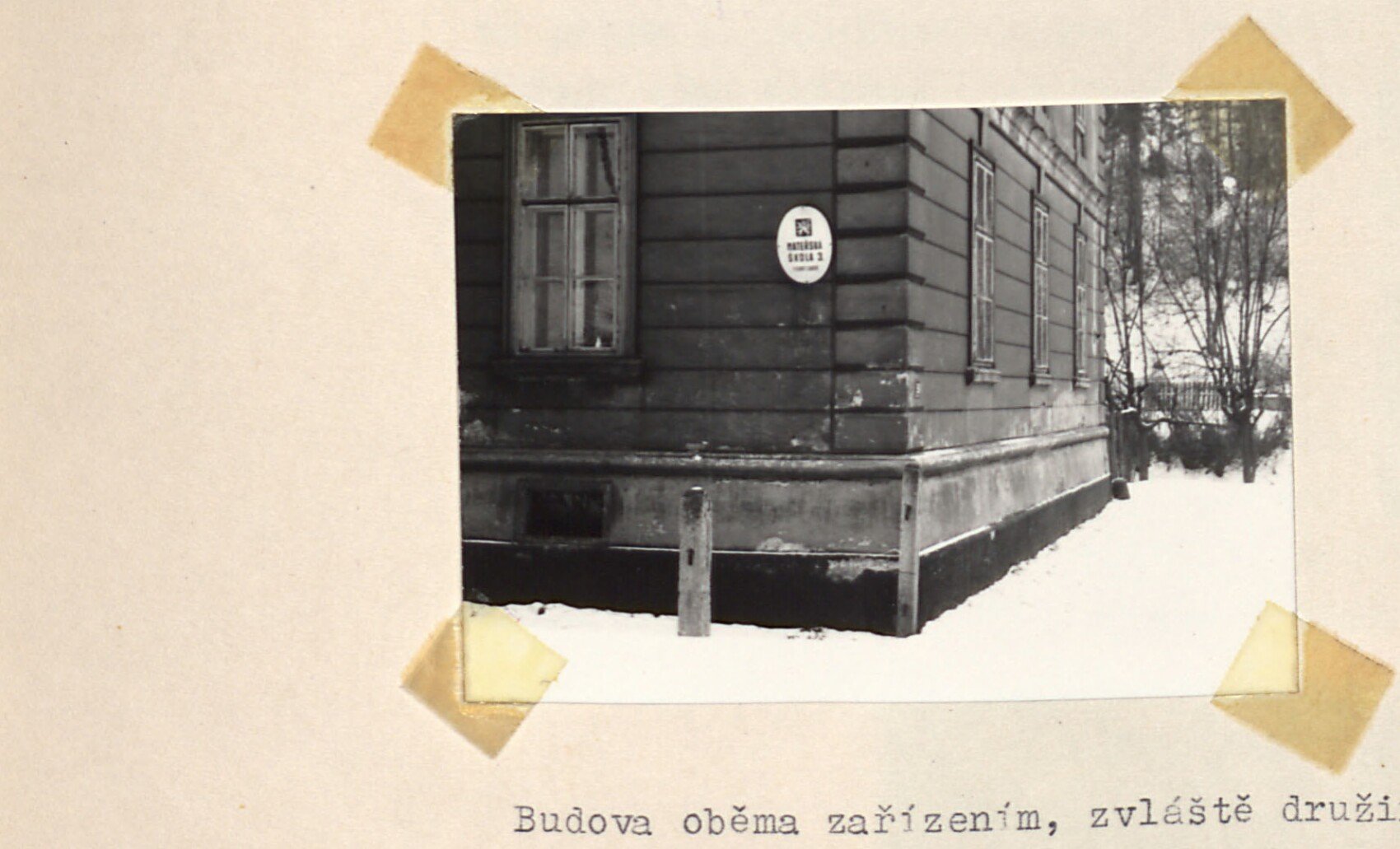 Dřevěný finský domek (roh budovy) na nízkých pilířích s informační cedulkou u okna — finský domek se školkou / družinou.