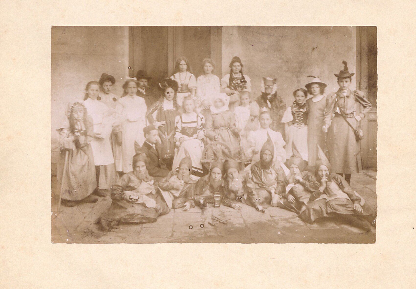 Sépiová skupinová fotografie z divadelního vystoupení školních dětí (Horní Slavkov, dle popisu z r. 1905). Početná skupina dětí a učitel v dobových kostýmech — dívky v historických šatech a kloboucích, postavy s čarodějnickými klobouky a vousy, vpředu leží převlečené děti (zřejmě trpaslíci) — patrně pohádková scéna. Vzadu zděná stěna se sloupem.