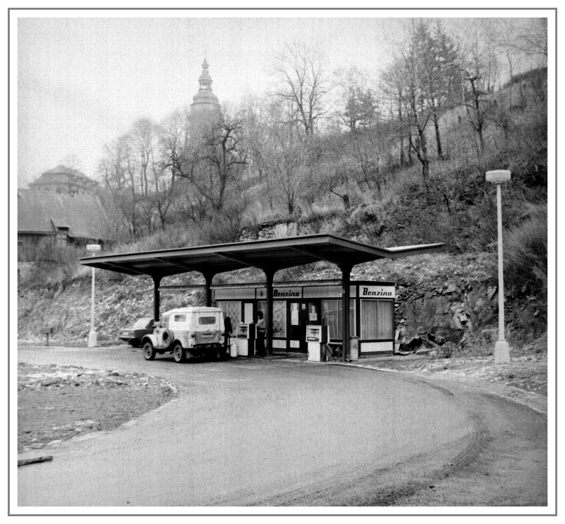 Čerpací stanice Benzina s přístřeškem  Horní Slavkov (Schlaggenwaldu) V pozadí nad svahem s náletovými dřevinami vyčnívá věž kostela