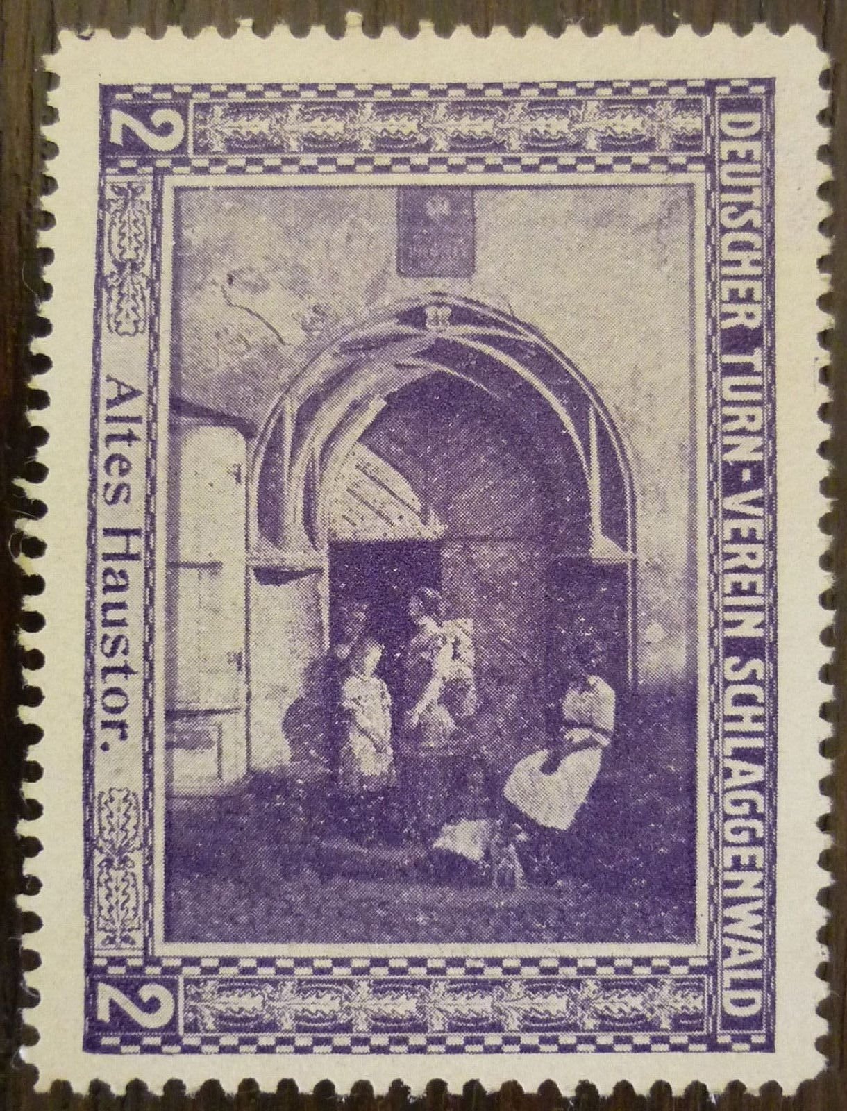 Reprodukce dobové (pravděpodobně charitativní) známky/nálepky spolku „Deutscher Turn-Verein Schlaggenwald" (Německý tělocvičný spolek Slavkov) s hodnotou „2" a vyobrazením brány s nápisem „Altes Haustor" (Stará domovní brána) – na obrázku klenutý portál, před ním dvě postavy v dobovém oděvu. Text na snímku: „DEUTSCHER TURN-VEREIN SCHLAGGENWALD" / „Altes Haustor." / „2"