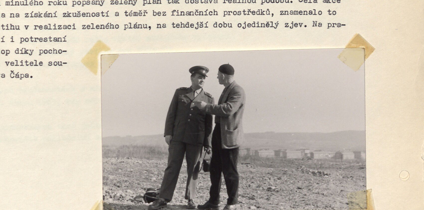 Černobílá fotografie dvou mužů hovořících na haldě/poli (skládka u šachty 3): vlevo muž v uniformě (zástupce velitele tábora Prokop), vpravo muž v civilu, v pozadí skladové buňky.