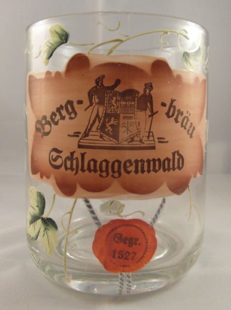 Studiová fotografie skleněného půllitru (pivního korbelu) s nálepkou „Berg-bräu Schlaggenwald" (Horní Slavkov) zdobenou motivem chmelových listů a reliéfním erbem se dvěma horníky; na skle je nalepena pečeť s nápisem „Gegr. 1527" (založeno 1527) odkazující na založení pivovaru, nikoli na stáří snímku. Fotografie na bílém pozadí má charakter moderní produktové/sběratelské fotografie.
