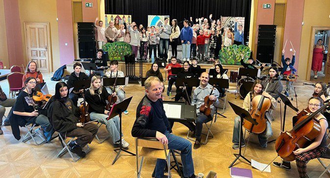 Orchestr ZUŠ band a pěvecké sbory při zkoušce/vystoupení v sále (Slavkovský zpravodaj, 6/2024)