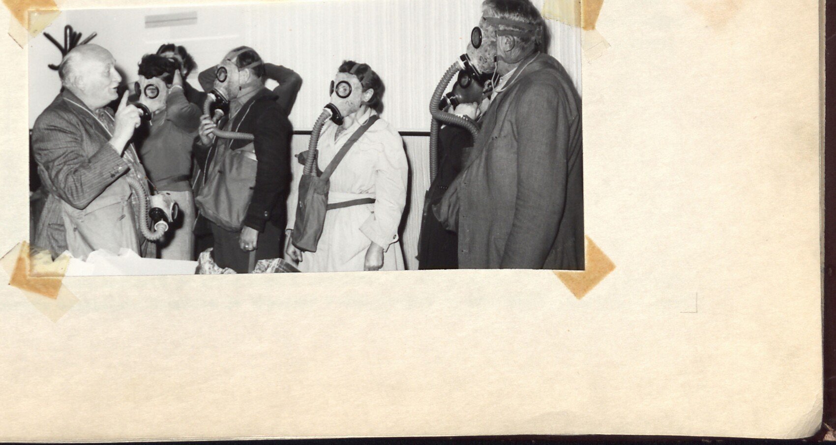 Černobílá fotografie skupiny občanů v plynových maskách při školení civilní obrany (CO); muži a ženy s maskami a vaky, vlevo starší muž v civilu.