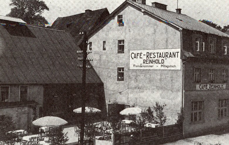 Pohlednicový snímek domu s nápisem „CAFÉ-RESTAURANT „REINHOLD"" a „Fremdenzimmer - Mittagstisch" na štítové zdi, na boční fasádě dále nápis „CAFÉ REINHOLD". Před domem zahrádka se slunečníky a stolky, vpravo dvoupatrová zástavba, vlevo hospodářské stavení se sedlovou střechou a elektrický sloup s izolátory.