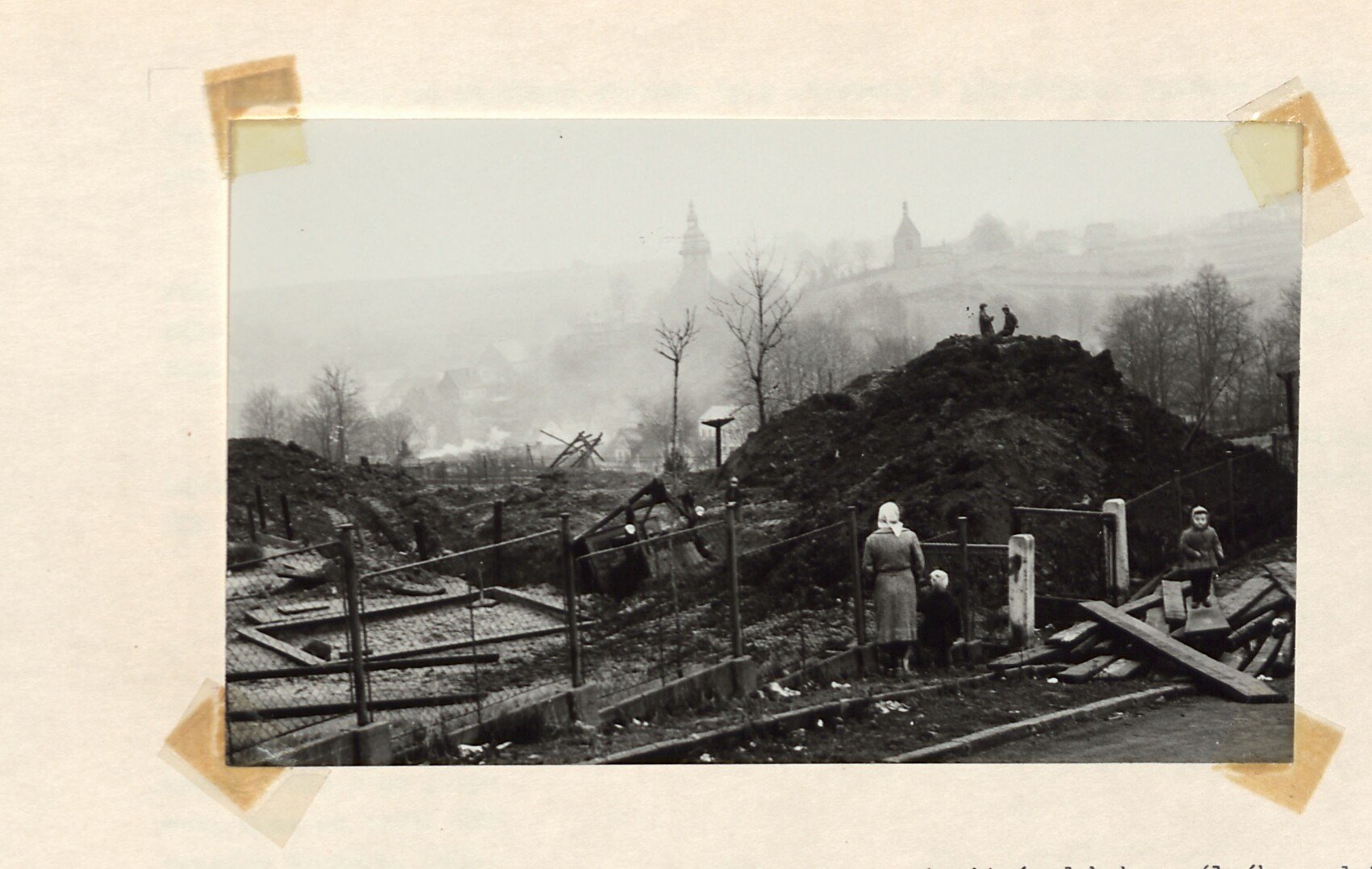 Černobílá fotografie: rozbořená, vyklízená část starého města – hromady hlíny a sutě, trosky dřevěných staveb, drátěný plot; v popředí stojí dvě ženy s dítětem, vzadu v mlze město s věžemi kostela. Asanace (demolice) staré zástavby v Horním Slavkově.
