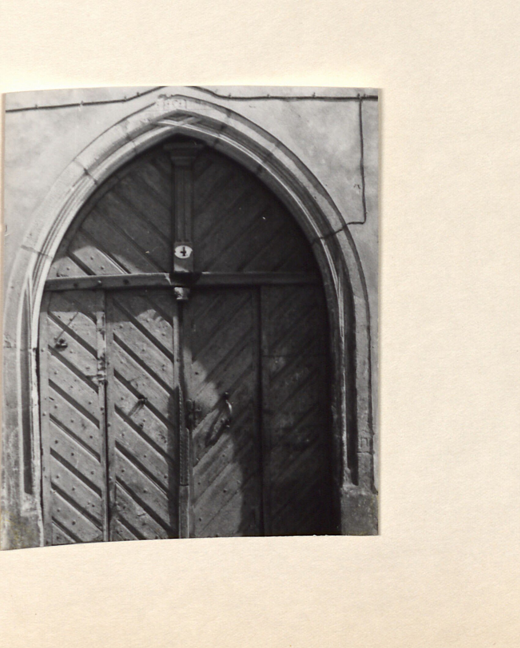Černobílá fotografie samostatně nalepená na pravém okraji strany – gotický lomený portál (vstupní dveře historické budovy) s dřevěnými vraty s kovaným kováním a zámkem.