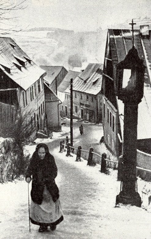 Zasněžená strmá ulička se schodištěm (Kirchstaffel) v Horním Slavkově, v popředí stará žena v dlouhém kabátu a šátku opřená o hůl, vpravo litinový sloup pouliční lampy zakončený křížem, v pozadí štítové domy a kostelní věž.
