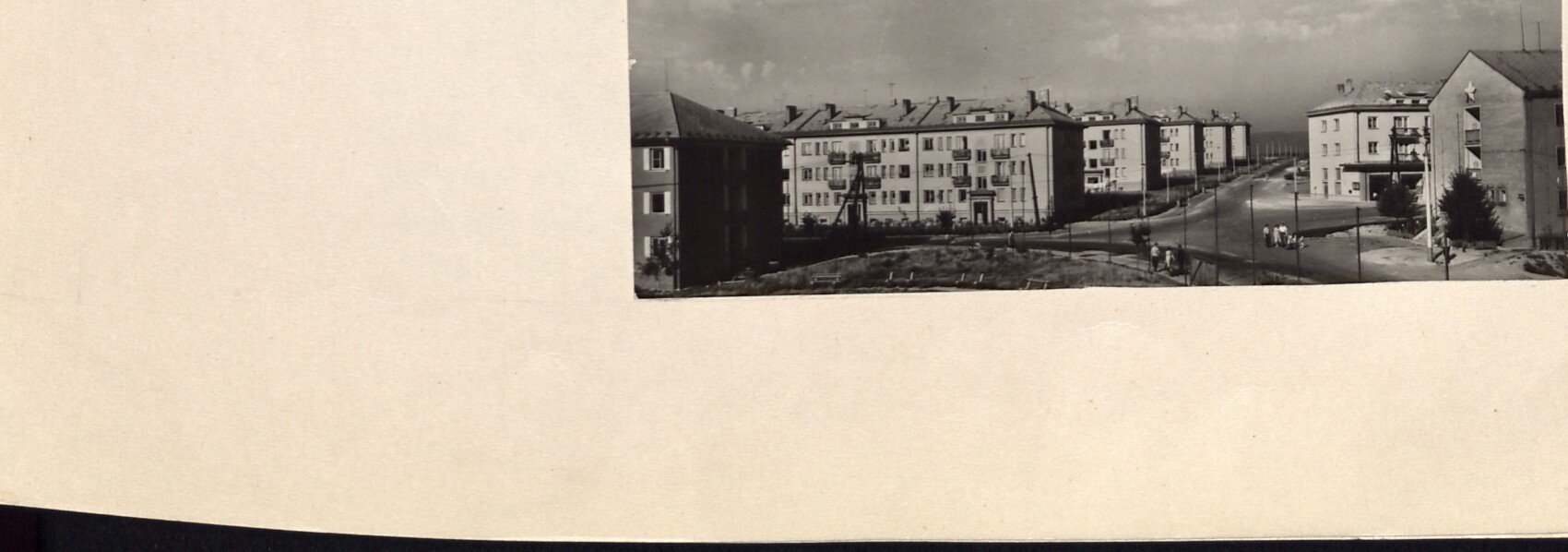 Černobílá fotografie: nově postavené zděné bytové domy sídliště s ulicí a lidmi; v pozadí krajina. Bez tištěné popisky.