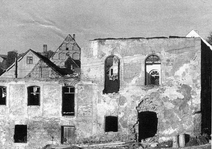 Demolice budovy č.207 v Horním Slavkově — fasáda s vylámanými okny a dveřmi, v pozadí vpravo štít hrázděného domu, vlevo přízemní domek se sedlovou střechou.