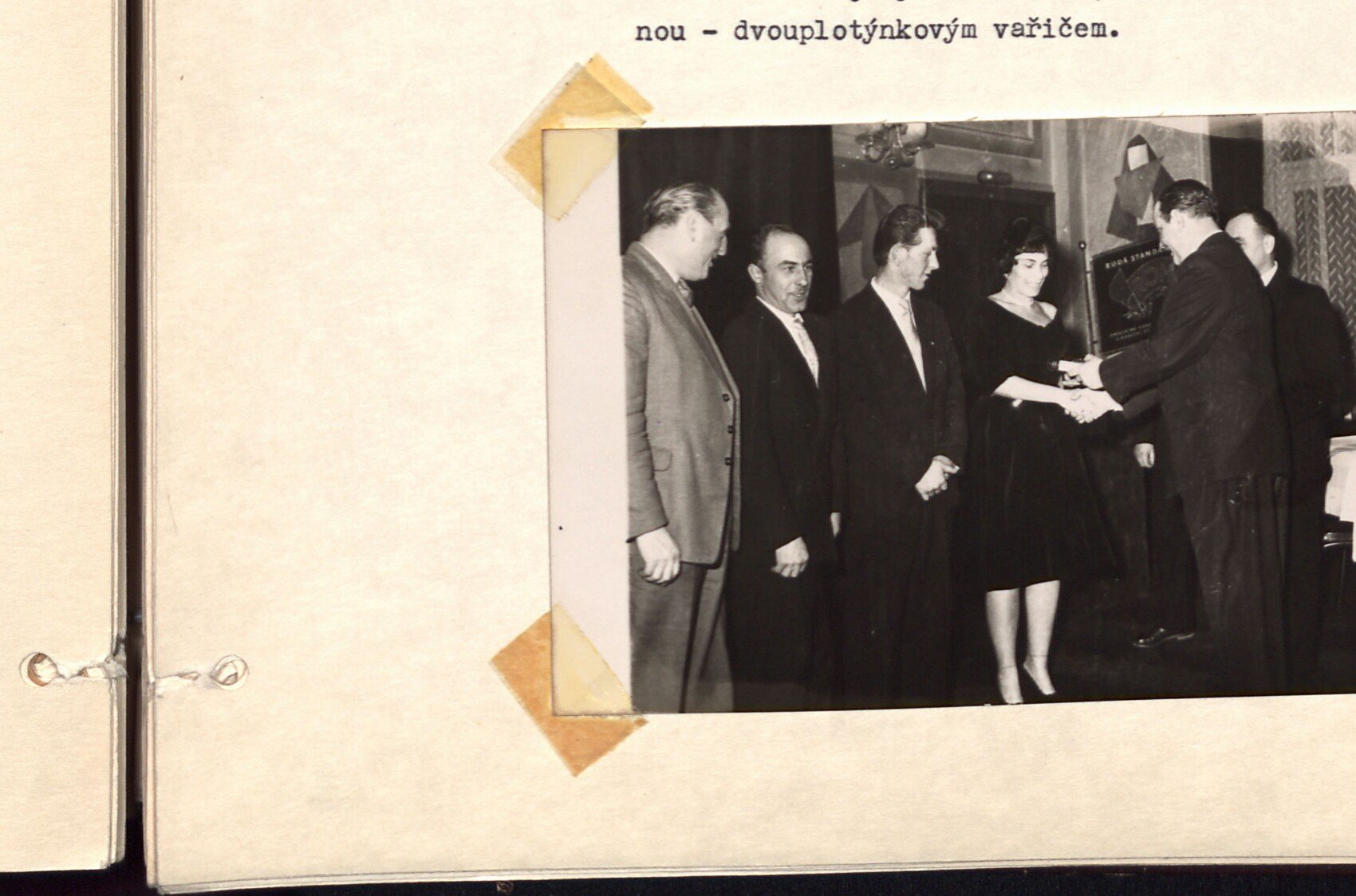 Slavnostní / společenské shromáždění — řada mužů v oblecích stojí vedle sebe, žena (vpravo) si podává ruku s mužem (předávání ocenění / poděkování). Černobílá fotografie v levé dolní části pravé strany.