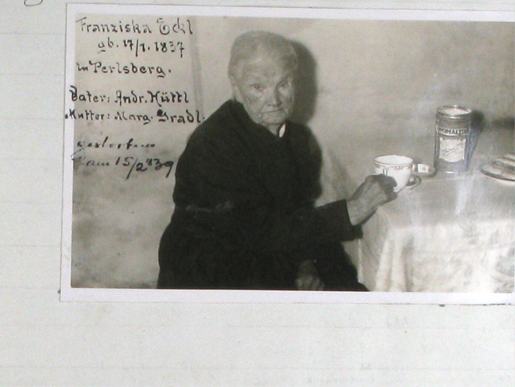Černobílá fotografie sedící stoleté ženy Franzisky Öckl u stolu, drží v ruce šálek; na stole je plechovka (zřejmě sladového nápoje). Rukopisná popiska na fotografii: „Franziska Öckl, nar. 17. 1. 1837 v Perlsbergu. Otec: Andr. Hüttl, matka: Marg. Bradl. Zemřela 15. 2. [19]39.“