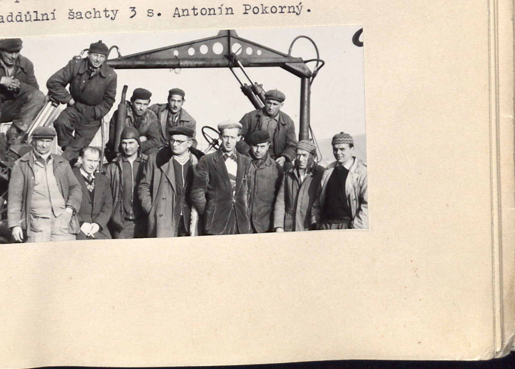 Skupinové foto mužů (dělníci / pracovníci) pózujících před strojem, patrně těžním zařízením nebo jeřábem; několik mužů sedí nahoře, ostatní stojí v řadě.