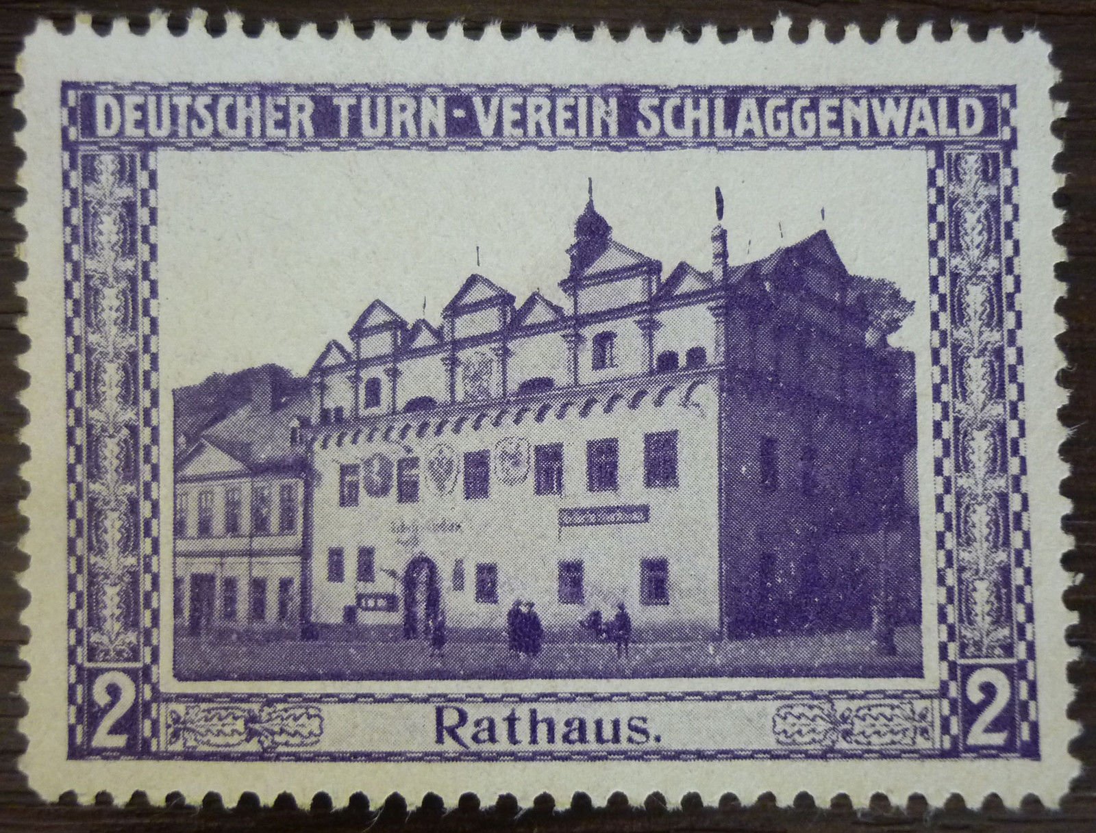 Dobročinná/reklamní známka (vinětka) Německého tělocvičného spolku Schlaggenwald s vyobrazením radnice (Rathaus) na náměstí, v popředí několik kolemjdoucích v dobovém oblečení. Nahoře nápis „DEUTSCHER TURN-VEREIN SCHLAGGENWALD", dole „Rathaus.", po stranách hodnota „2".
