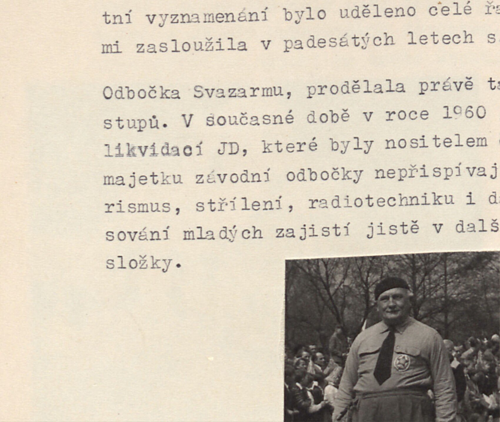 Muž v baretu s odznakem (rozhodčí?) vede ovčáckého psa, za ním dav diváků.