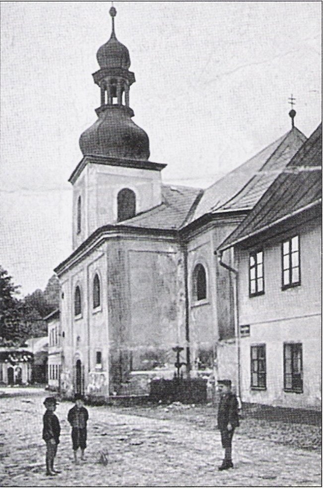 Kostel sv. Anny (podle názvu souboru „sv.Anna") s cibulovitou věží, pohled od dlážděné ulice s přilehlou zástavbou. V popředí na dlážděné ulici dva chlapci a muž v dobovém oblečení (klobouky, dlouhé kabáty).