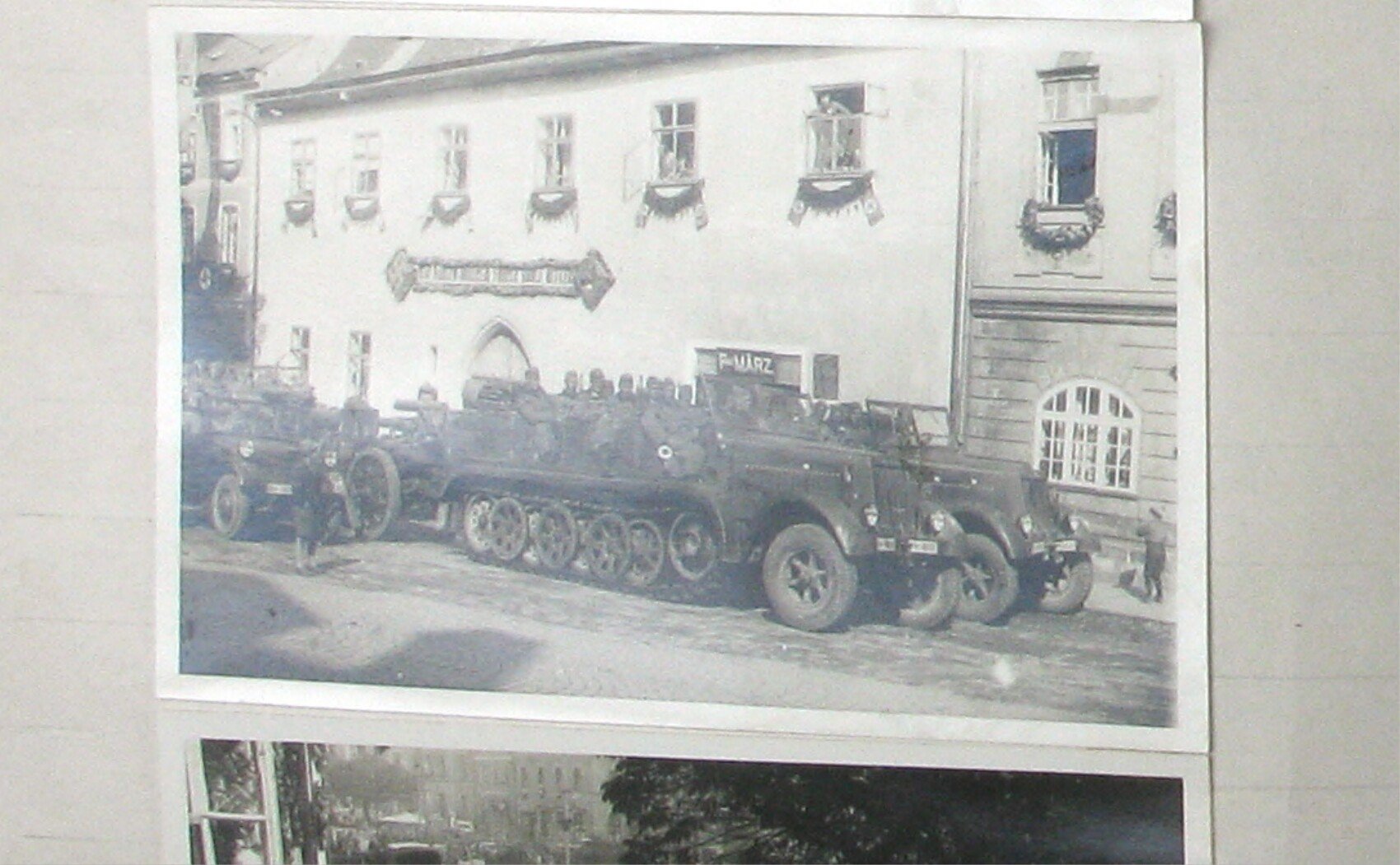 Kolona obrněných polopásových vozidel a tažených děl stojící podél velké budovy (hostinec/dům s vývěsním štítem) ozdobené girlandami a prapory.