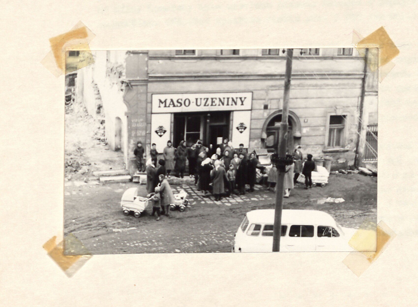 Černobílá fotografie: fronta lidí (ženy s taškami, dětským kočárkem a nákupními koši) před obchodem s nápisem "MASO-UZENINY" v Horním Slavkově; před obchodem zaparkovaná dodávka/auto, vlevo trosky zbořeného domu. Ilustruje zásobovací potíže roku 1962.